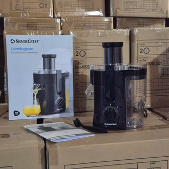 A SilverCrest countertop centrifugal juicer in black and clear plastic, shown next to its original retail box and a small instruction booklet on top of stacked cardboard shipping boxes.
